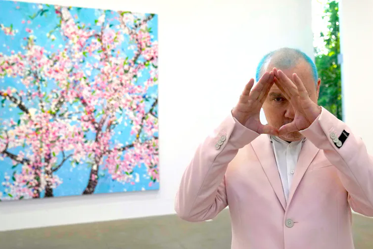 Blue-haired artist Damien Hirst in a pink suit poses in front of one of his Cherry Blossoms artworks.