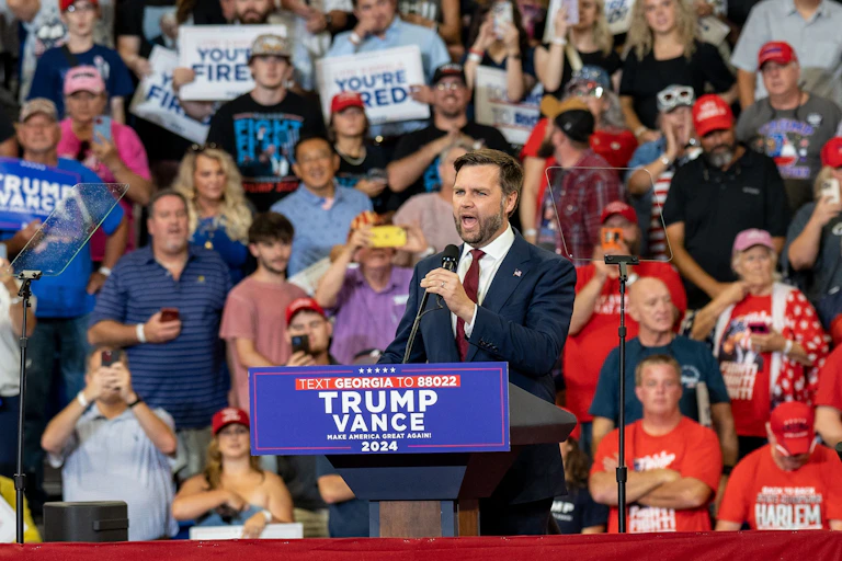J. D. Vance da un discurso en una tarima con varios seguidores a sus espaldas.