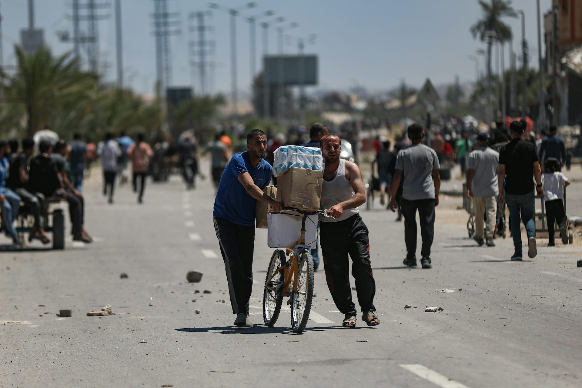 Two men push a bicycle loaded with aid supplies while others walk towards an aid distribution point.