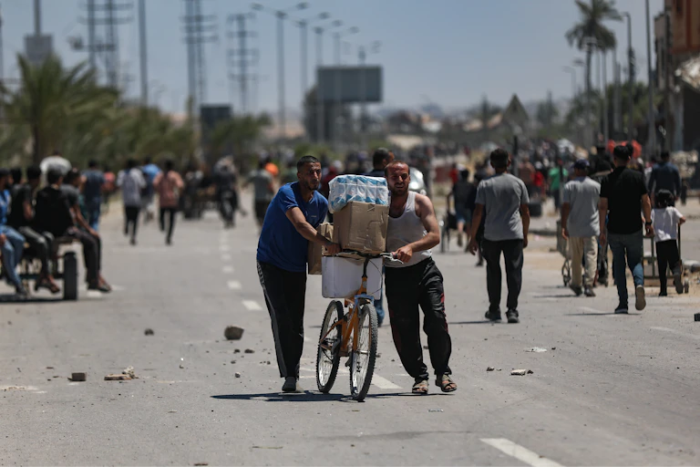 Two men push a bicycle loaded with aid supplies while others walk towards an aid distribution point.