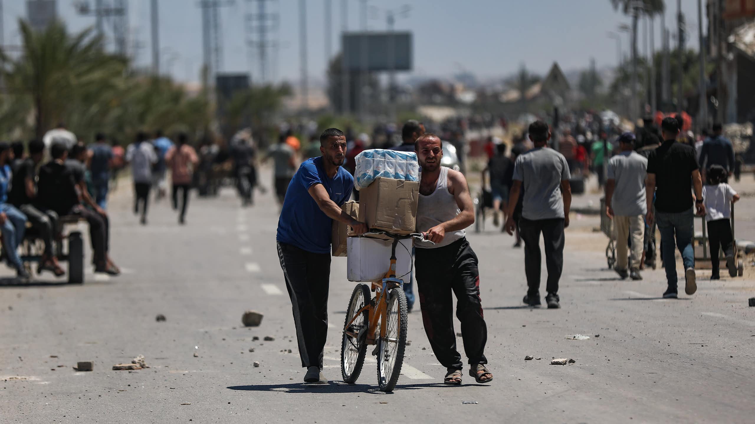 Two men push a bicycle loaded with aid supplies while others walk towards an aid distribution point.