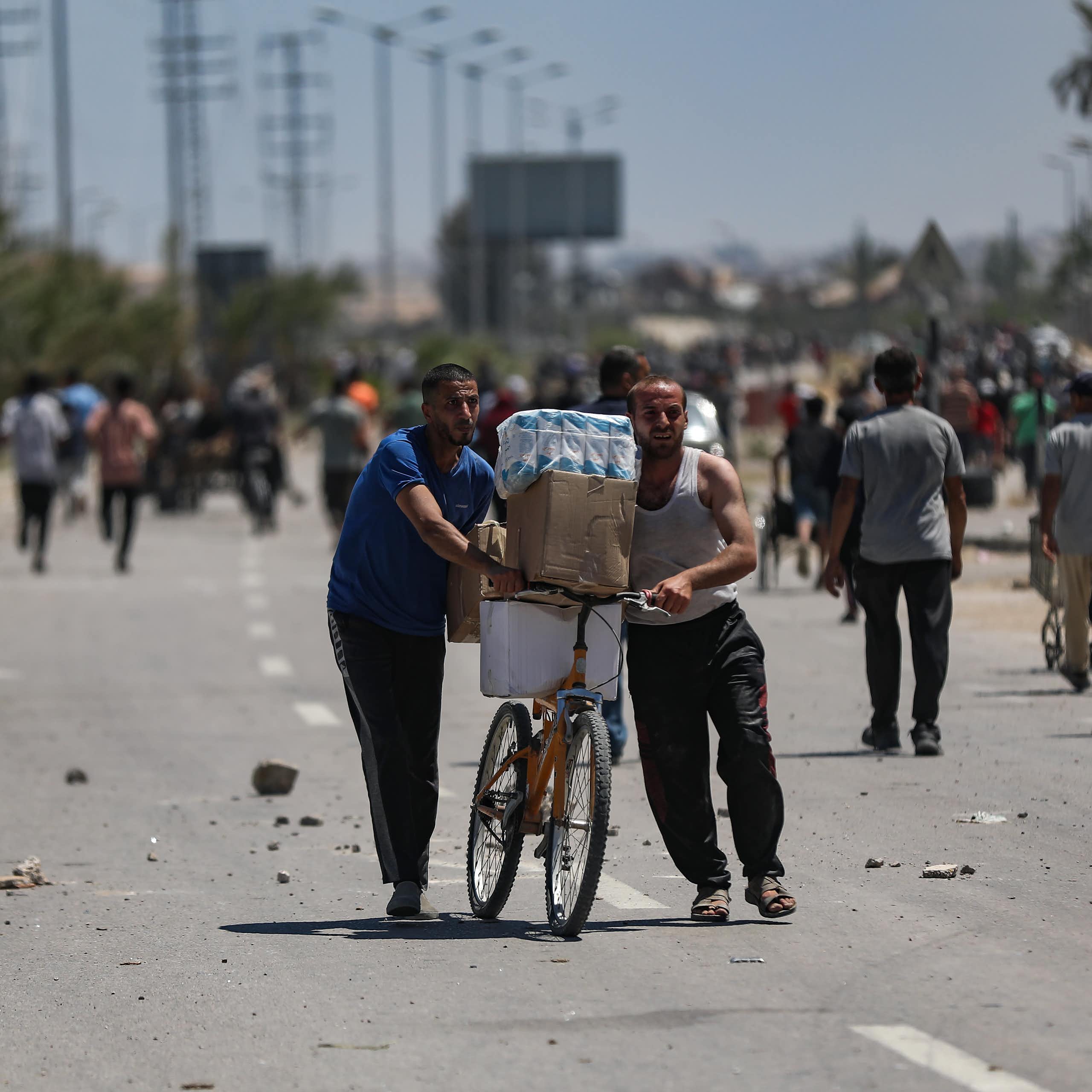 Two men push a bicycle loaded with aid supplies while others walk towards an aid distribution point.