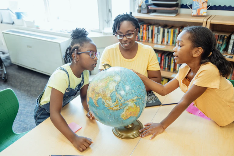 Two students and an adult wearing yellow shirts examine a globe while sitting at a table in a library.