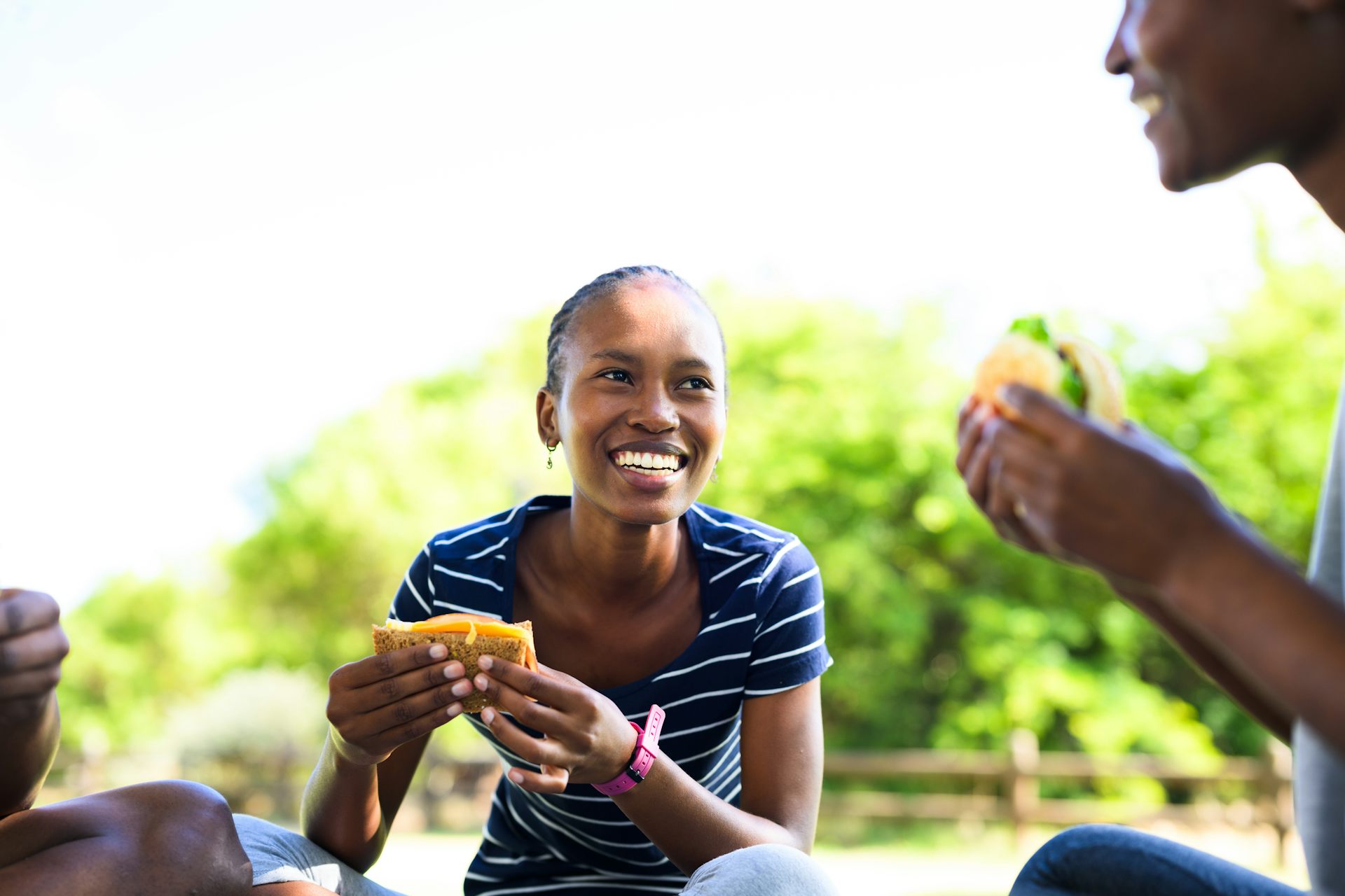 A young woman sitting on the grass outside eating a brown bread and cheese sandwich