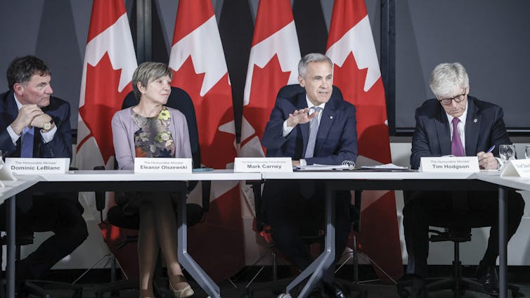 Un hombre con un cabello gris corto se encuentra en una mesa larga con dos hombres y mujeres más con una serie de banderas canadienses detrás de ellas.