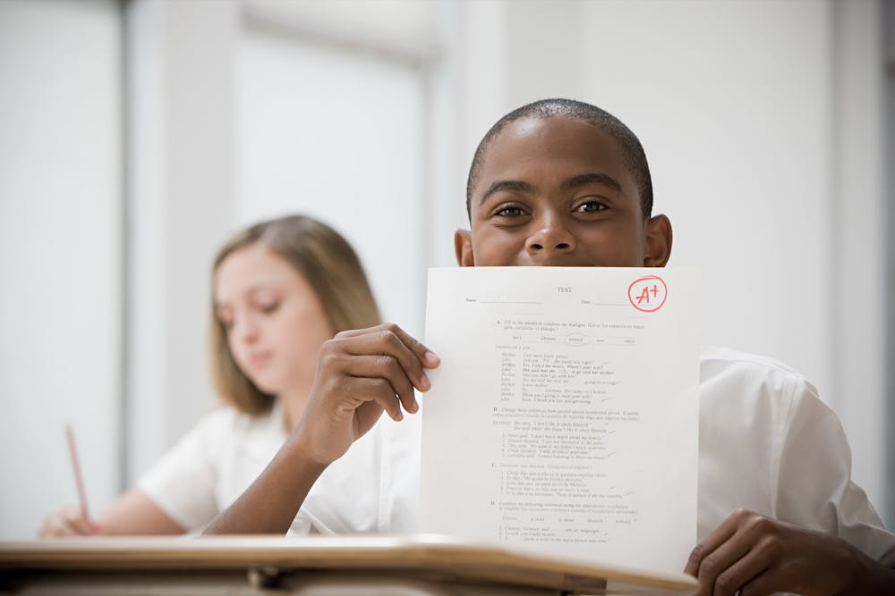 A boy wearing a white shirt holds up an exam paper with an A+ grade.