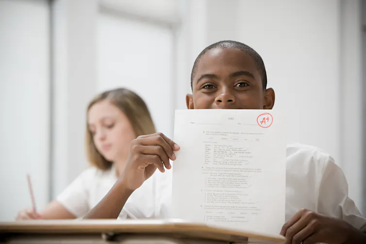A boy wearing a white shirt holds up an exam paper with an A+ grade.