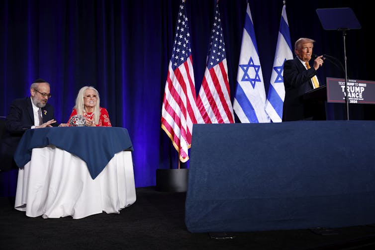 A man standing at a podium in front of US and Israeli flags, speaking to a gathering.