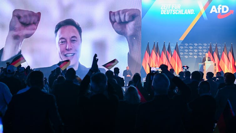 A smiling man raising both hands on a large screen in front of a big crowd.