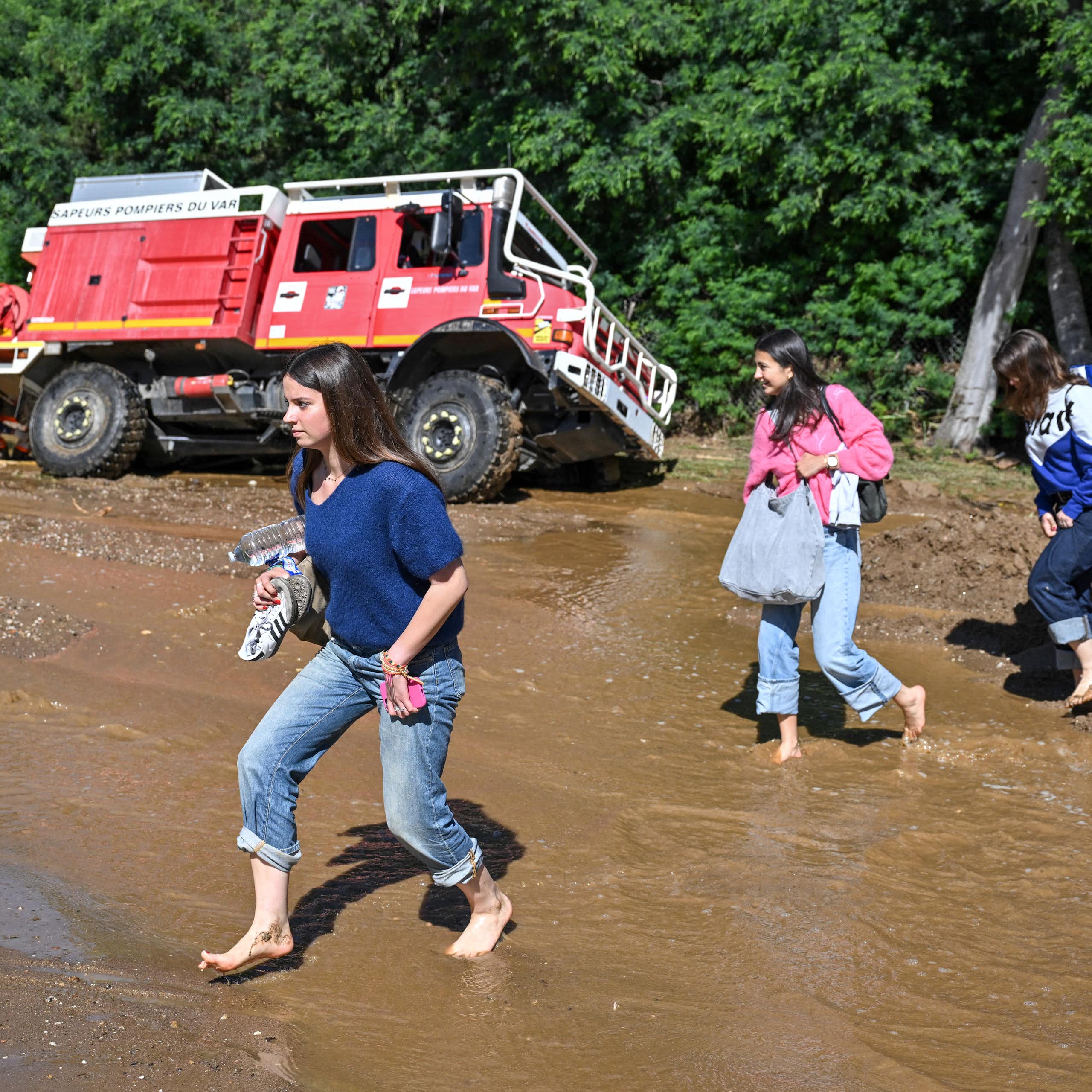 trois personnes marchent pied nus sur un sol inondé et boueux, à l'arrière un camion de pompier