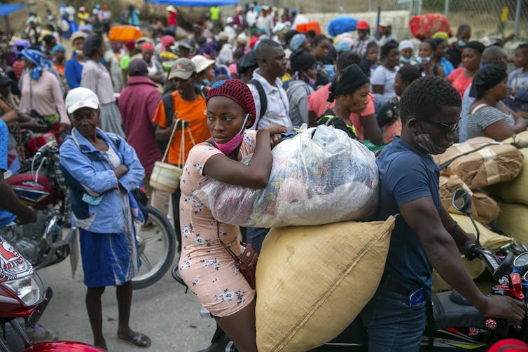 Para los migrantes haitianos en la República Dominicana, "la reproducción es como una pena de muerte" 3 La mujer viajero está sentada en una bicicleta apoyada en grandes bolsas. El conductor del hombre tiene manijas. Detrás de ellos una gran multitud