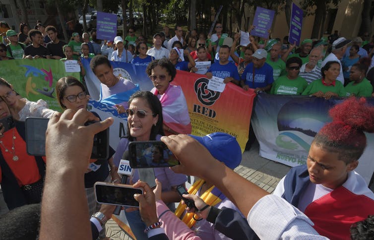 Para los migrantes haitianos en la República Dominicana, "la reproducción es como una pena de muerte" 2 Una mujer con gafas de sol habla a un grupo de personas que sostienen micrófonos y teléfonos, detrás de ella, las mujeres se encuentran con una gran señal