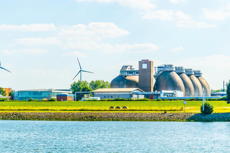 Vista de una planta industrial y aerogeneradores a orillas de un río.