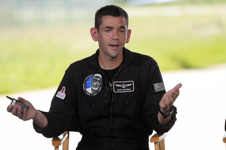 A man with dark hair and a black jumpsuit sitting in a chair and speaking.