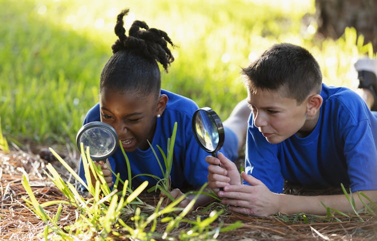 Two children wearing blue shirts lay on the grass with magnifying glasses, exploring the environment.