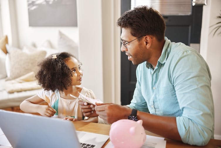 With a piggy bank in the foreground, a man holding money talks to a young girl about finances.