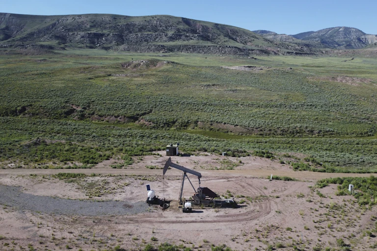 A pumpjack operates in an area of greenery with mountains as a backdrop.