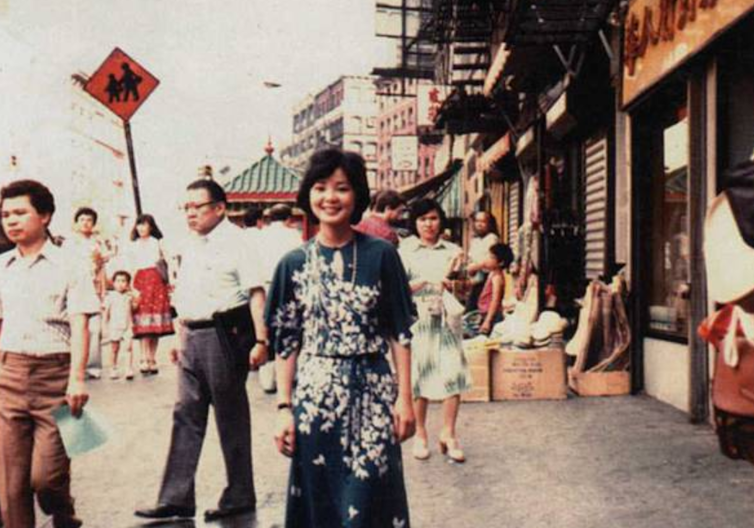 Young woman wearing blue dresse smiles and poses on a sidewalk filled with pedestrians.