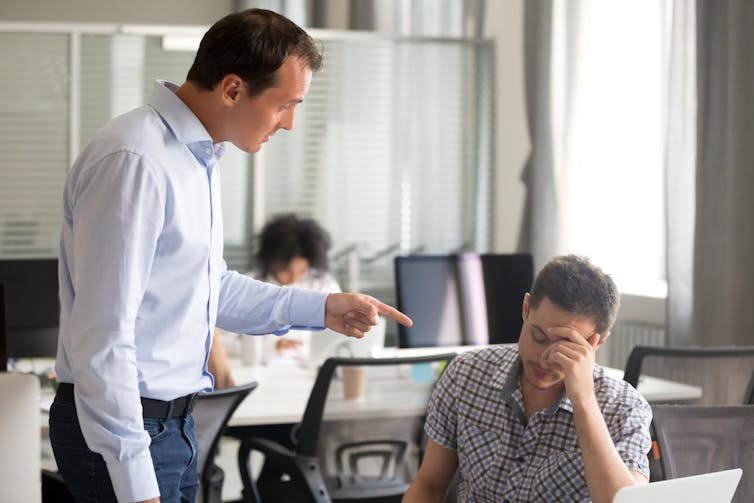 A man stands next to another man, who is seated at a desk, and points a finger at him while speaking forcefully