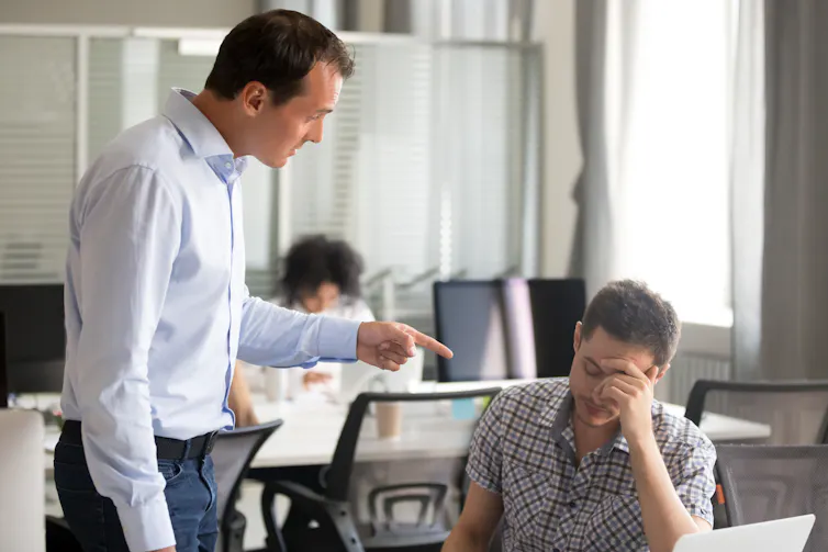A man stands next to another man, who is seated at a desk, and points a finger at him while speaking forcefully