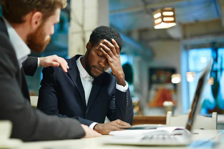A man rests his hand on the shoulder of another man who has an upset look on his face and is resting his head against his hand