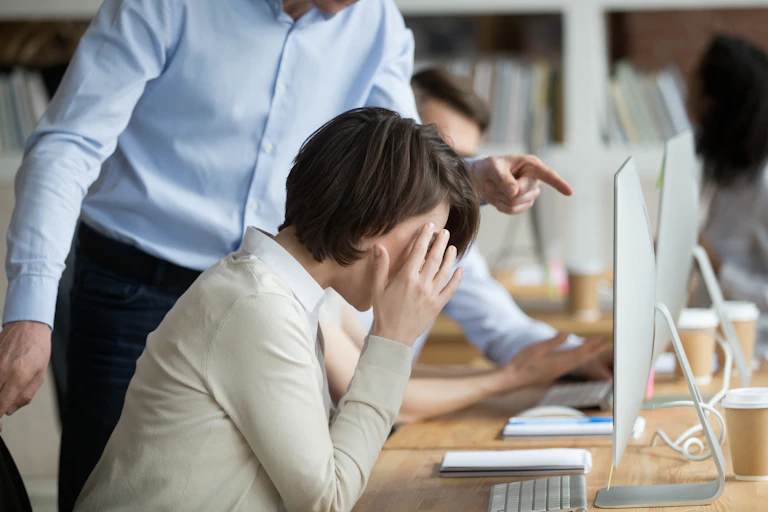 A person seen from the neck down looms over a woman who is sitting at a desk hiding her face in her hands