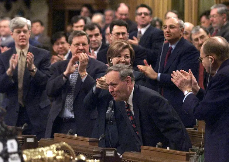 A man has a self-satisfied smile on his face as he stands up in the House of Commons, surrounded by people applauding him.