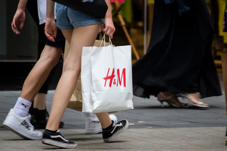 Two people seen from the waist down walking down the street, with one person carrying an H&M shopping bag