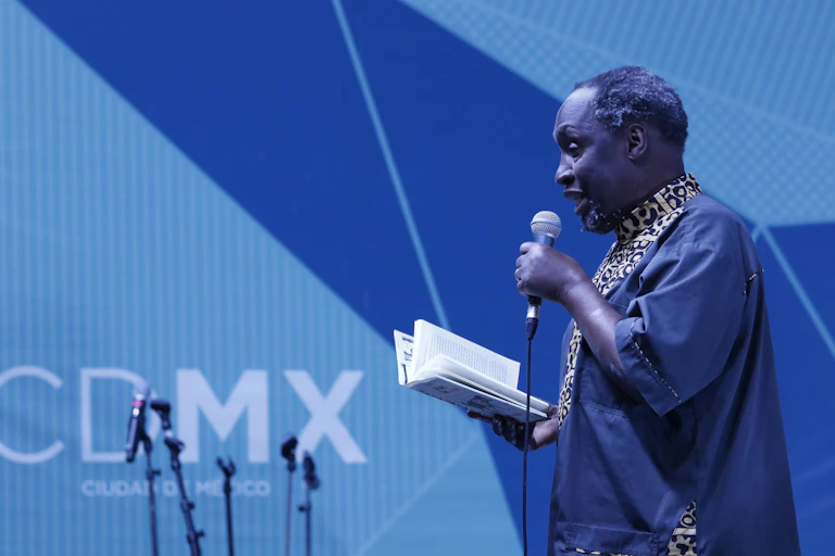 An older African man dressed in blue traditional attire on a stage against a blue backdrop. He holds a book in his one hand and a microphone in the other.