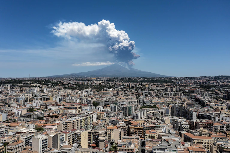 Un panache de cendres s'élève d'un volcan, avec une ville au premier plan.