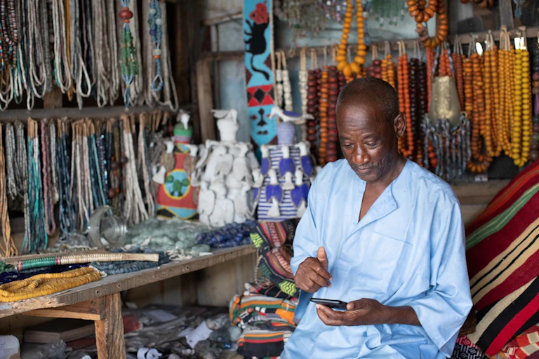 An older man sitting in a market stall, looking at a phone