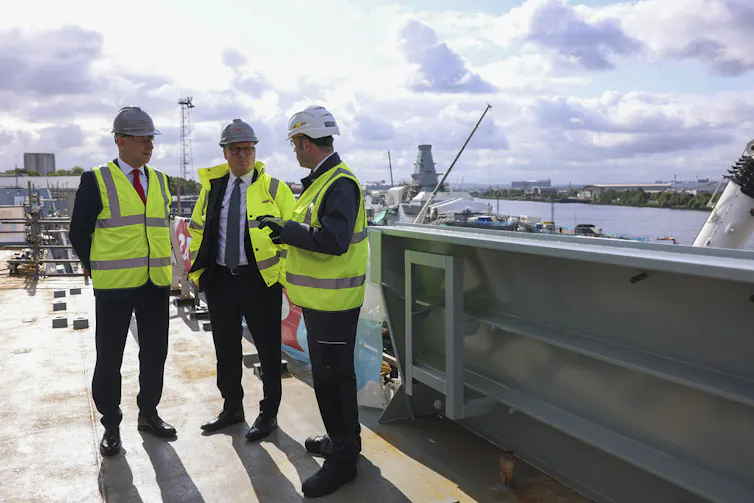 Starmer and Healey in high-vis vests and hard hats, speaking with a third man on the deck of a ship