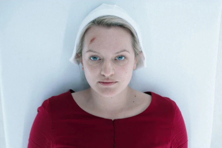 Elizabeth Moss lying on a medical table in a red dress and white cap.