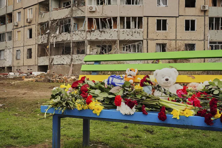 Toys and flowers on a playground bench with a damaged apartment building in the background.