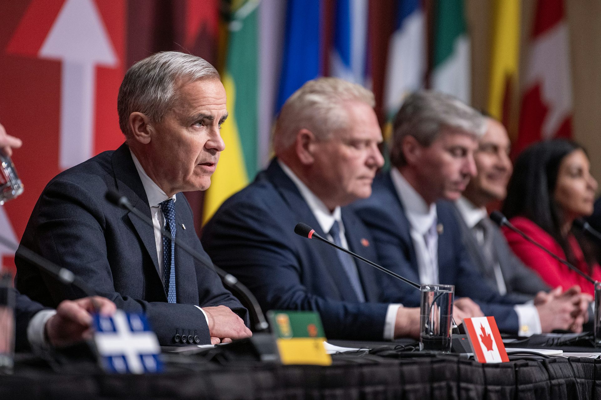 A man with short grey hair speaks; other men and a woman sit beside him at a long table with provincial/territorial flags behind them
