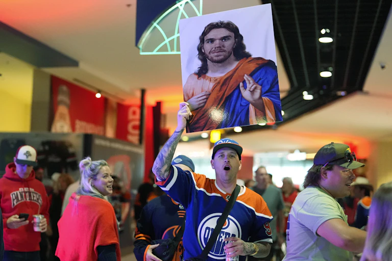 A man in a blue and orange jersey holds a large poster of a man in a Jesus-like pose, dressed in orange and blue robes.