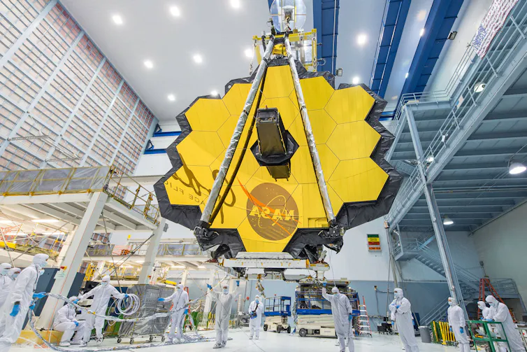 The JWST's mirror, which looks like a large, roughly hexagonal shiny surface made up of 18 smaller hexagons put together, sitting in a facility. The mirror is reflecting the NASA meatball logo.