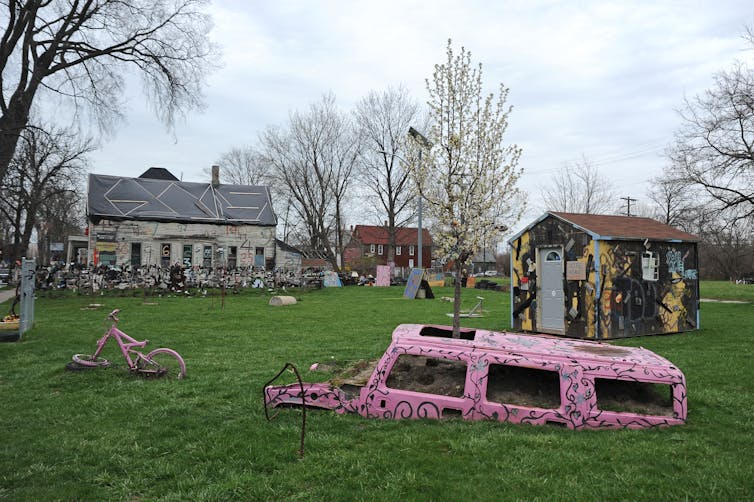 A pink bus is half buried in a field of green grass, and buildings in the distance are painted brightly.