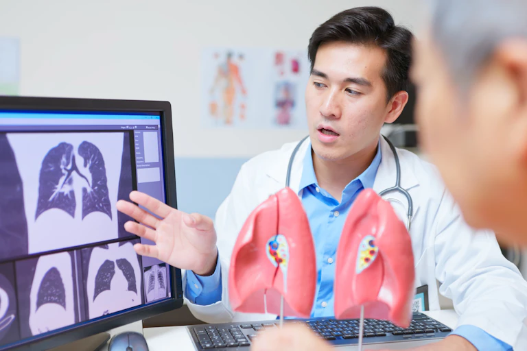 Doctor pointing to lung imaging scans on screen, models of lungs on table, older male patient looking at screen