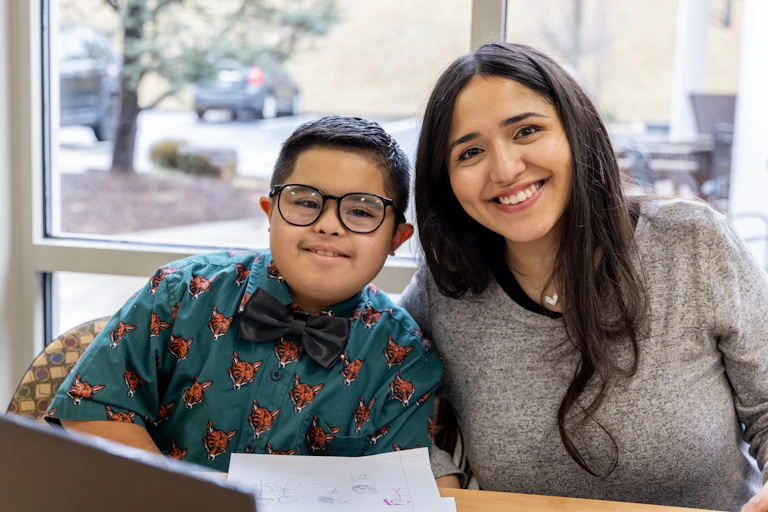 A student and teacher sitting side by side in a classroom lean in for a photo.