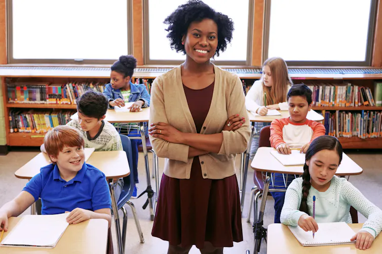 A Black teacher stands before rows of students sitting at desks.
