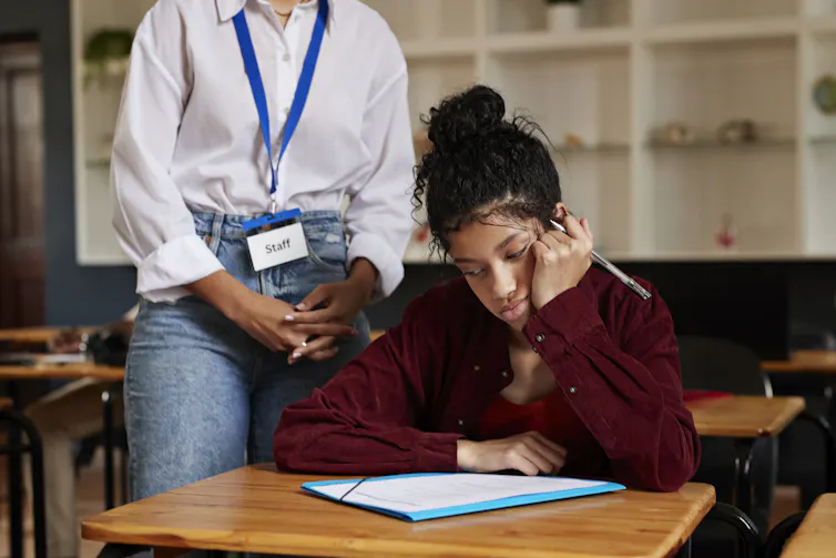 A frustrated student sits at a desk while a teacher stands behind her in the background.