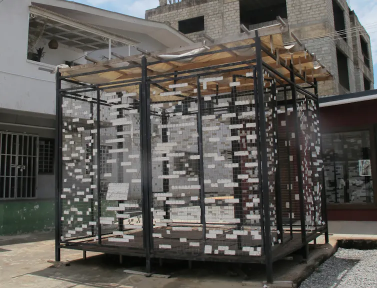 In an urban courtyard is a square structure with wire walls and hundreds of pieces of white paper.
