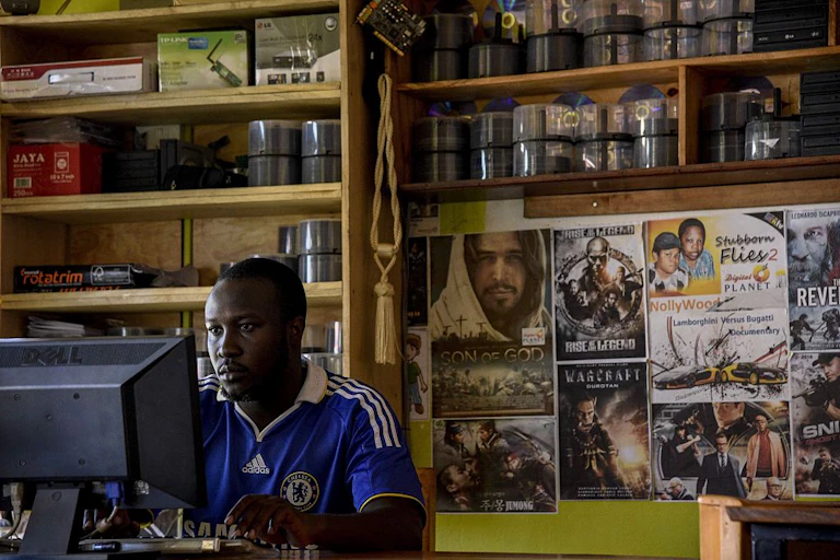 A man seated at a computer with shelves and posters on the wall behind him