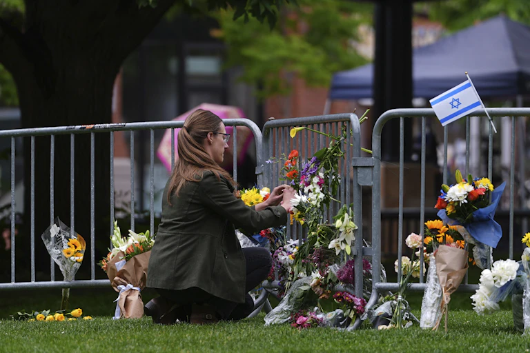 A woman kneels near a metal fence and places flowers among other bouquets and a small blue and white Israeli flag.