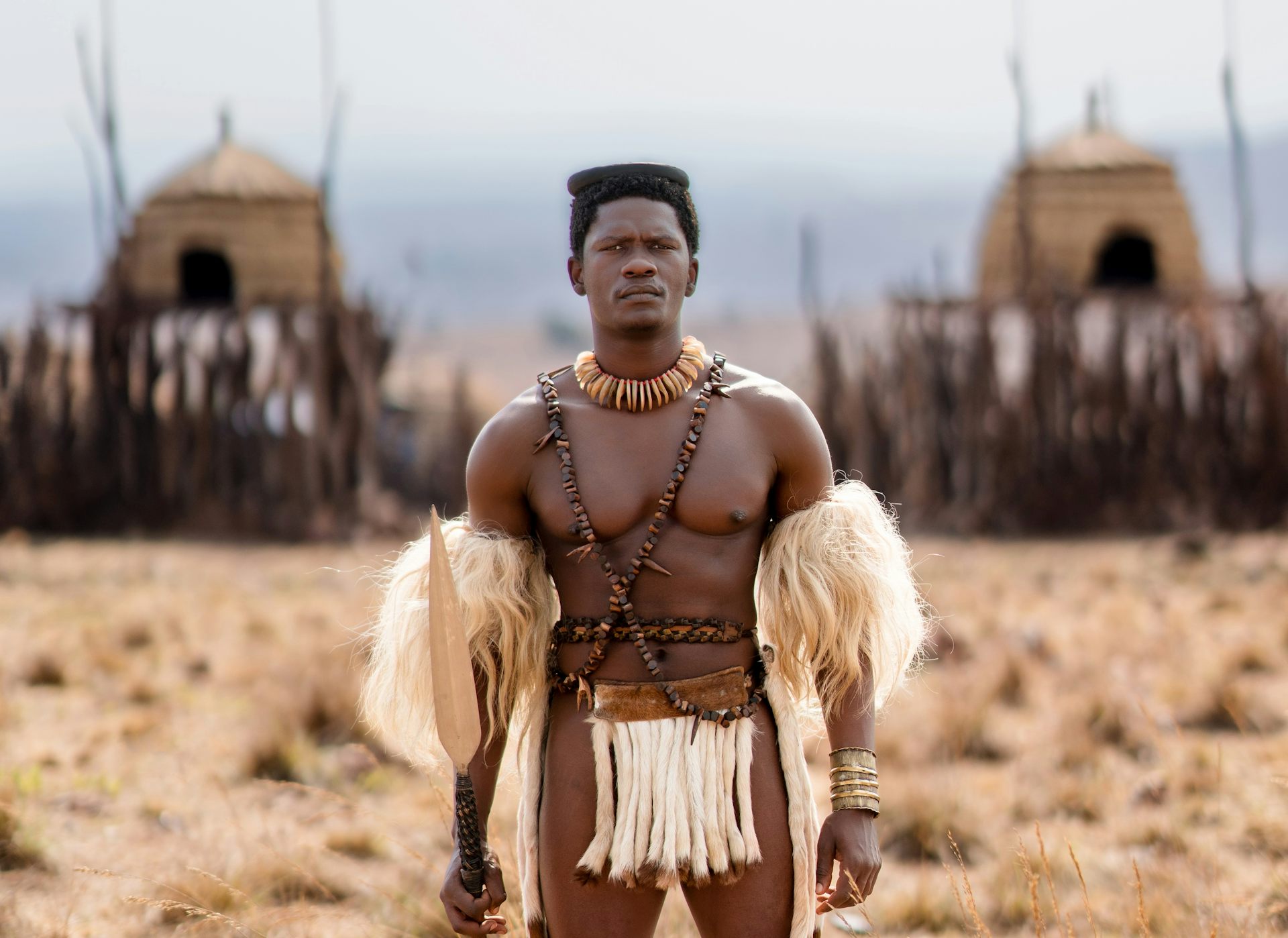 A muscular man stands in front of a reed and wood homestead wearing armbands and a codpiece of animal hair, beads across his chest.
