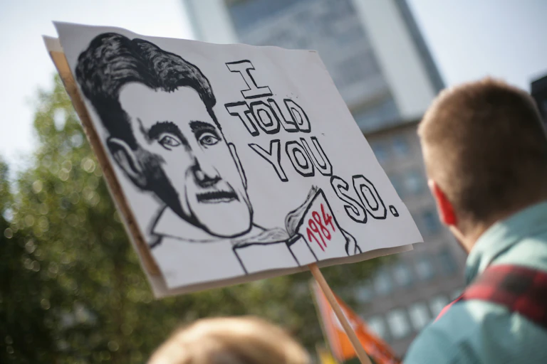 A sign being held by a man that says 'I told you so' and has a picture of George Orwell and his book 1984.