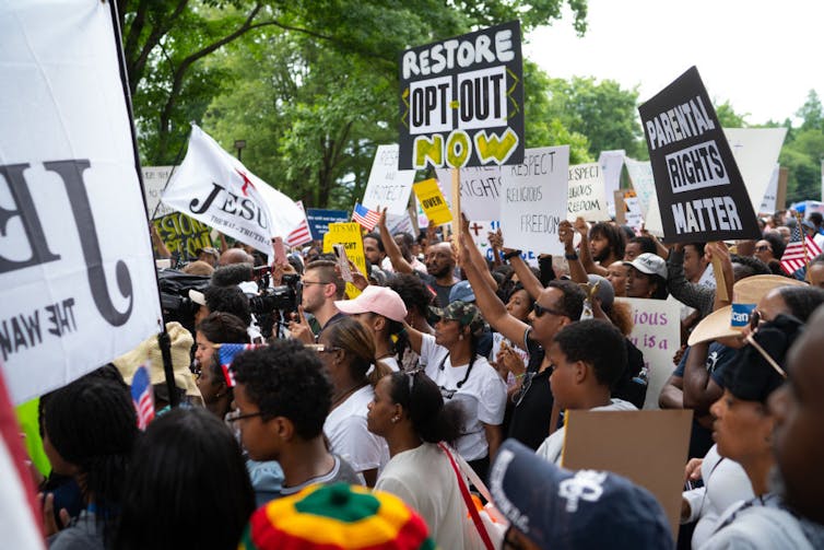 A large crowd protesting outside and holding black-and-white signs with slogans like 'Parental rights matter.'