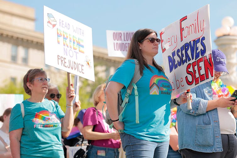 Several people, some of them wearing bright blue shirts with rainbows, hold signs outside a large stone building.