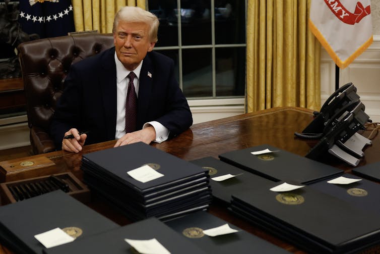 A light-haired man in a suit holding a pen at a desk covered with folders.